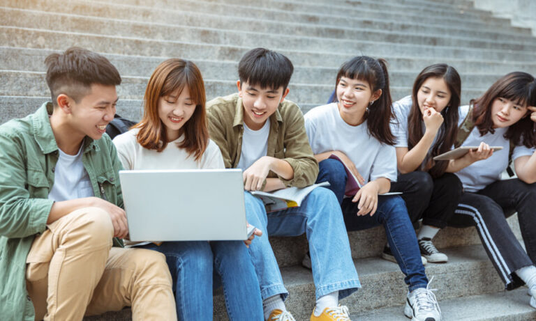 Asian students sitting in a row on concrete steps, using wireless devices